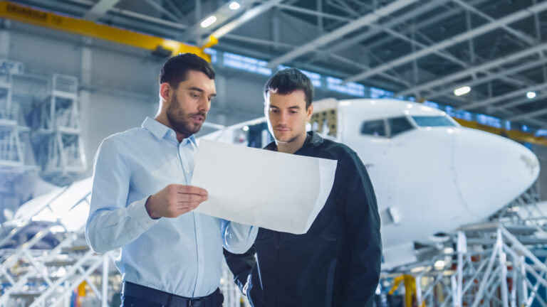 Aircraft Maintenance Worker and Engineer having Conversation. Holding Project Blueprint